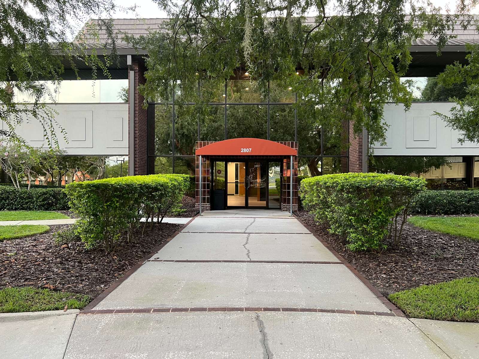 The front walk to our building at 2807 W Busch Blvd: a hedge-lined path to glass doors under a red awning, beneath a canopy of oaks